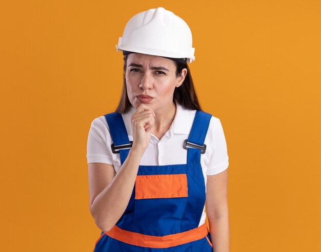 Mujer joven constructora sospechosa en uniforme agarró la barbilla aislada en la pared naranja