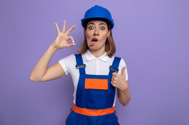 Foto gratuita mujer joven constructora en uniforme de construcción y casco de seguridad confundido haciendo el signo de ok mostrando el pulgar hacia arriba de pie en púrpura