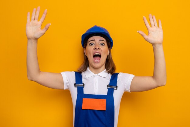 Mujer joven constructora en uniforme de construcción y casco de seguridad conmocionada y asustada levantando los brazos en pánico de pie sobre la pared naranja
