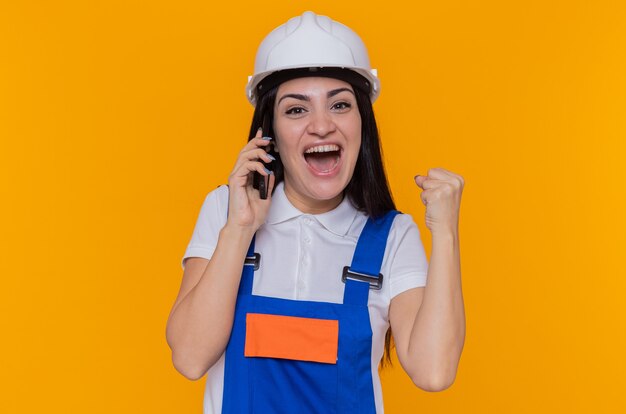 Mujer joven constructora en uniforme de construcción y casco de seguridad feliz y emocionado apretando el puño mientras habla por teléfono móvil de pie sobre la pared naranja