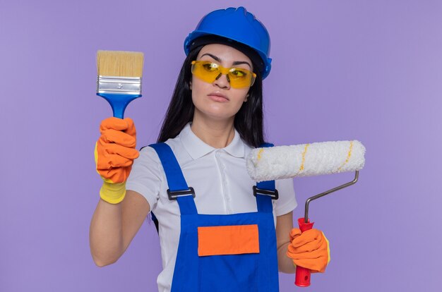 Mujer joven constructora en uniforme de construcción y casco de seguridad con guantes de goma sosteniendo rodillo de pintura y pincel mirándolo con cara seria de pie sobre la pared púrpura