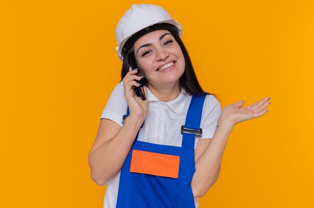 Mujer joven constructora en uniforme de construcción y casco de seguridad mirando al frente sonriendo levantando el brazo mientras habla por teléfono móvil de pie sobre la pared naranja