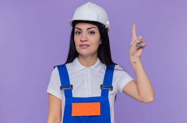 Mujer joven constructora en uniforme de construcción y casco de seguridad mirando al frente con una sonrisa en la cara inteligente que muestra el dedo índice con una gran idea nueva de pie sobre la pared púrpura