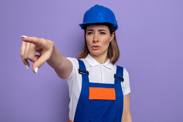 Mujer joven constructora en uniforme de construcción y casco de seguridad mirando a un lado con cara seria apuntando con el dedo índice a algo de pie en púrpura