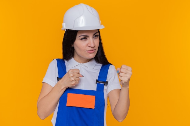 Mujer joven constructora en uniforme de construcción y casco de seguridad mirando a un lado con expresión seria y segura con el puño cerrado como un luchador de pie sobre una pared naranja