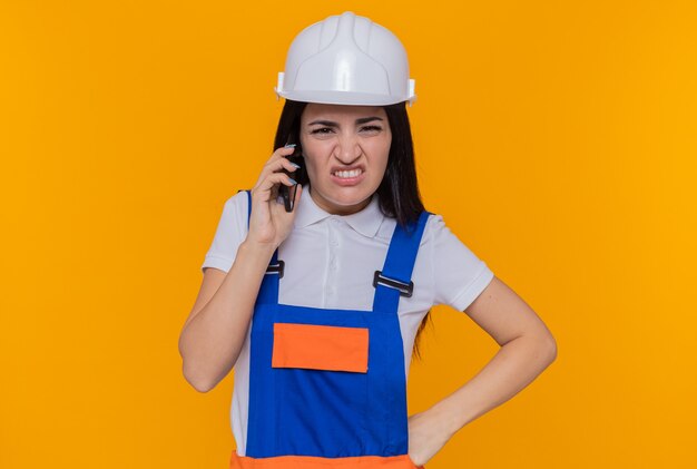 Mujer joven constructora en uniforme de construcción y casco de seguridad mirando molesto y disgustado mientras habla por teléfono móvil de pie sobre una pared naranja