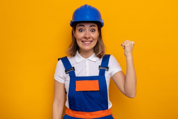 Mujer joven constructora en uniforme de construcción y casco de seguridad que parece feliz y emocionada levantando el puño como un ganador