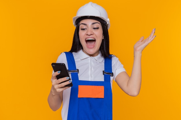 Mujer joven constructora en uniforme de construcción y casco de seguridad con smartphone mirándolo feliz y emocionado con el brazo levantado de pie sobre la pared naranja