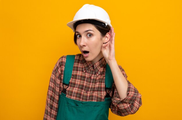 Mujer joven constructora en uniforme de construcción y casco de seguridad sorprendido sosteniendo la mano en la oreja tratando de escuchar chismes de pie en naranja