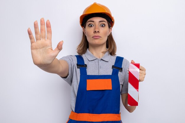 Mujer joven constructora en uniforme de construcción y casco de seguridad sosteniendo cinta adhesiva preocupada haciendo gesto de parada con la mano de pie sobre la pared blanca