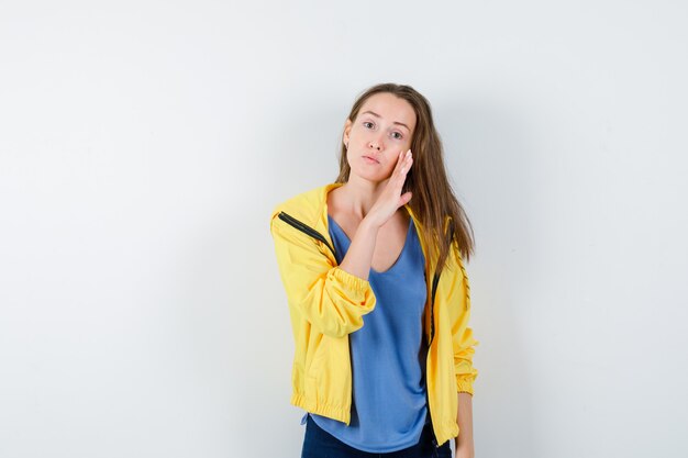 Mujer joven contando secretos detrás de la mano en camiseta, chaqueta y mirando preocupado, vista frontal.