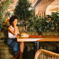 Foto gratuita mujer joven contemplada sentada en la mesa del restaurante con tableta digital; celular y agenda