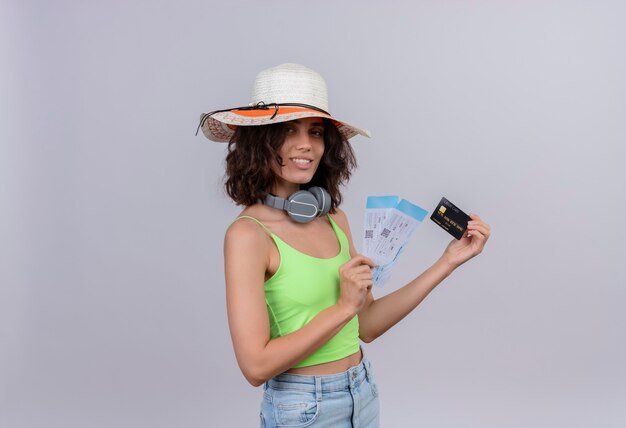 Una mujer joven contenta con el pelo corto en verde crop top con sombrero para el sol mostrando billetes de avión y tarjeta de crédito sobre un fondo blanco.