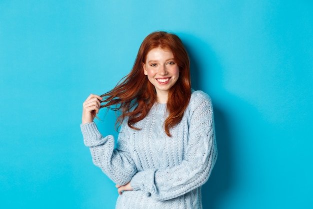 Mujer joven coqueta con el pelo rojo, jugando con el pelo y sonriendo, de pie en suéter sobre fondo azul.