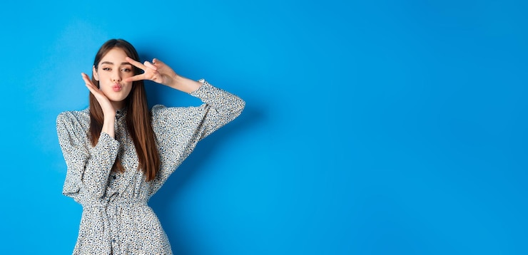 Foto gratuito mujer joven coqueta en vestido mostrando vsign en el ojo y labios fruncidos para besar de pie feliz en azul