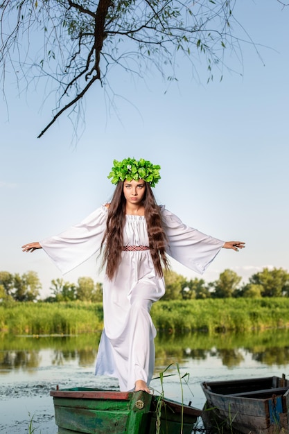 Mujer joven con corona de flores en la cabeza, relajándose en un barco en el río al atardecer. Hermoso cuerpo y cara. Fotografía de arte fantástico. Concepto de belleza femenina, descanso en el pueblo.