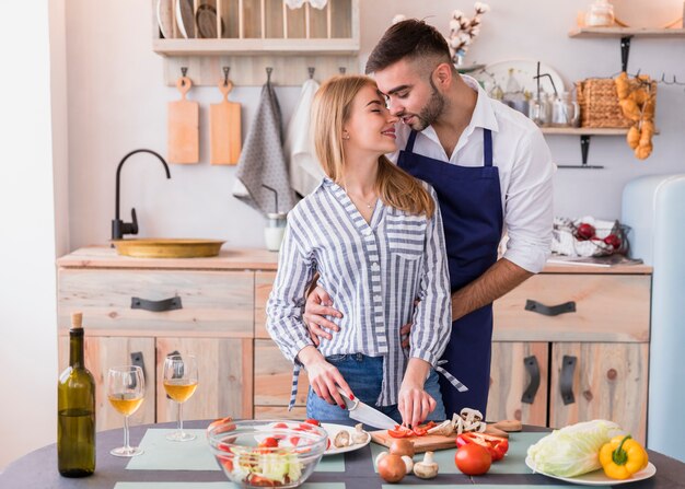 Mujer joven cortando verduras mientras que hombre abrazándola