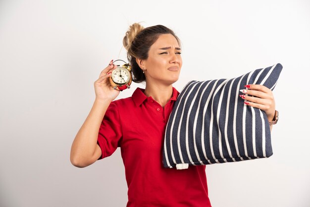 Foto gratuita mujer joven en corto rojo mostrando un despertador y sosteniendo una almohada.