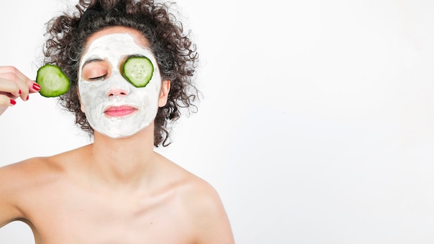 Mujer joven con cosméticos en la cara que aplica el pepino a su ojo