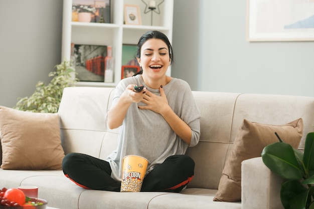 Mujer joven con cubo de palomitas de maíz sosteniendo el control remoto del televisor a la cámara sentado en el sofá detrás de la mesa de café en la sala de estar