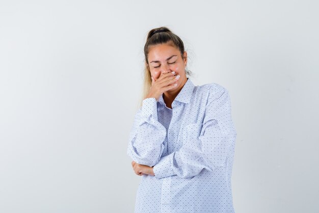 Mujer joven cubriendo la boca con la mano, sonriendo, manteniendo los ojos cerrados con camisa blanca y mirando feliz