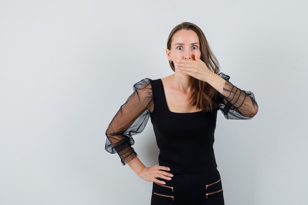 Mujer joven cubriendo la boca con una mano y sosteniendo la otra mano en la cintura con blusa negra y pantalón negro y mirando sorprendido. vista frontal.