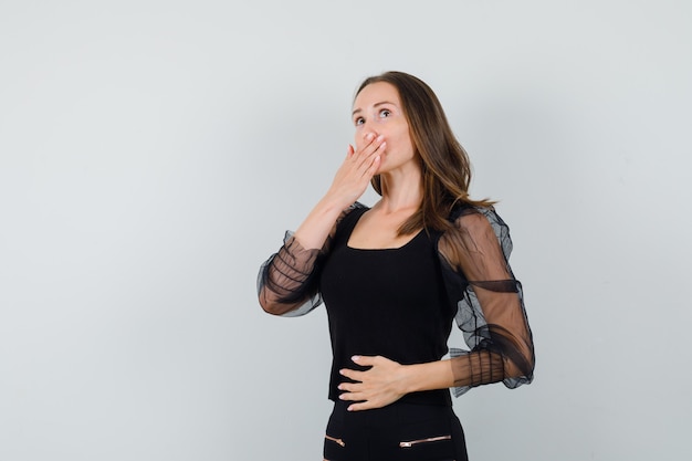 Mujer joven cubriendo la boca con una mano y sosteniendo la otra mano en el vientre con blusa negra y pantalón negro y mirando tímida. vista frontal.