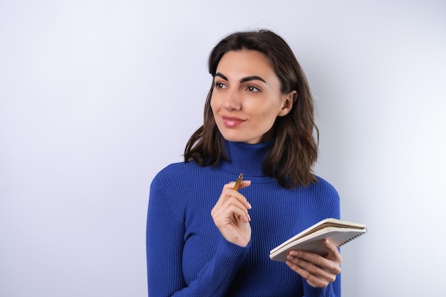 Mujer joven en un cuello alto de golf azul sobre un fondo blanco pensativo con un cuaderno en la mano piensa en ideas metas para el año