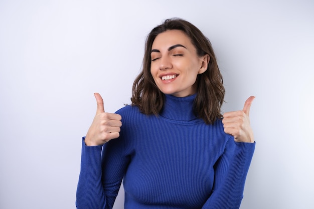 Mujer joven en un cuello alto de golf azul sobre un fondo blanco con una sonrisa segura sonriendo alegremente mostrando los pulgares hacia arriba