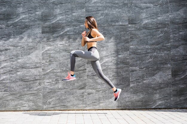 Mujer joven con cuerpo en forma saltando y corriendo contra la pared gris.
