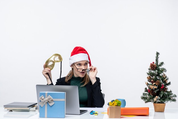 Mujer joven curiosa con gafas de sombrero de santa claus y máscara sentado en una mesa con un árbol de Navidad y un regalo en la oficina sobre fondo blanco.