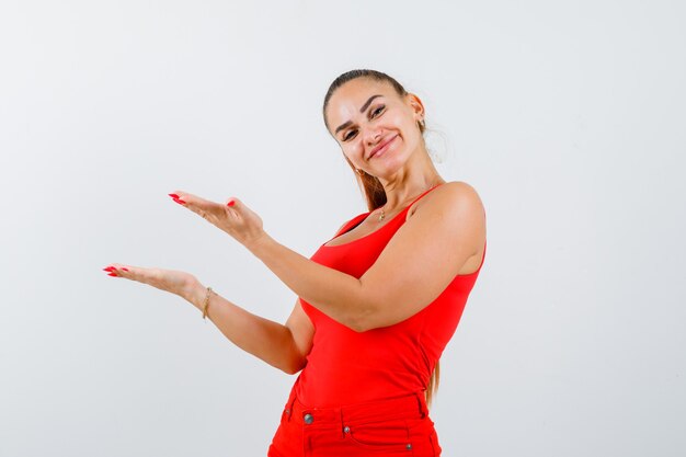 Mujer joven dando la bienvenida a algo en camiseta roja sin mangas, pantalones y luciendo bonita, vista frontal.