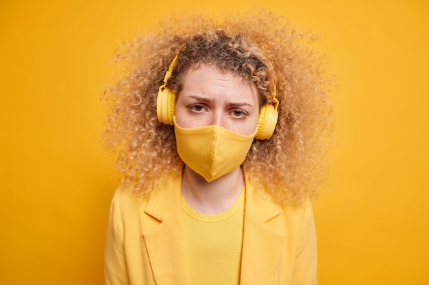 Mujer joven decepcionada y disgustada que usa una máscara protectora se siente sola en casa sola en autoaislamiento escucha música a través de auriculares estéreo inalámbricos posa contra la pared amarilla