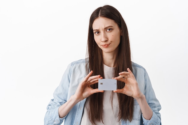 Mujer joven decepcionada que muestra la tarjeta de crédito del banco, sonríe y se ve dudosa, frunciendo el ceño disgustado, de pie sobre una pared blanca