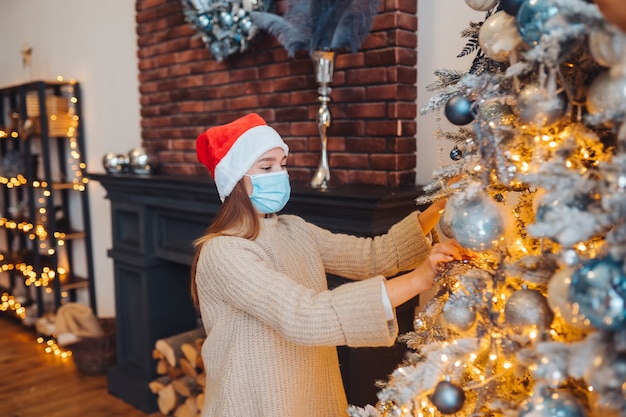 Una mujer joven decora el árbol de Navidad con máscara médica