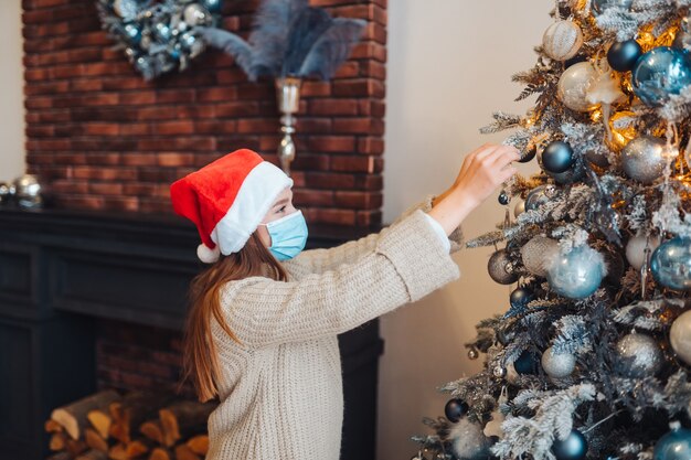 Una mujer joven decora el árbol de Navidad con máscara médica