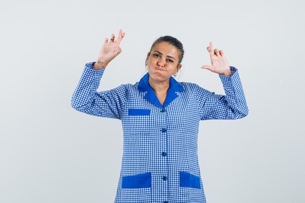 Mujer joven con los dedos cruzados, hinchando las mejillas en camisa de pijama de cuadros azules y luciendo bonita. vista frontal.