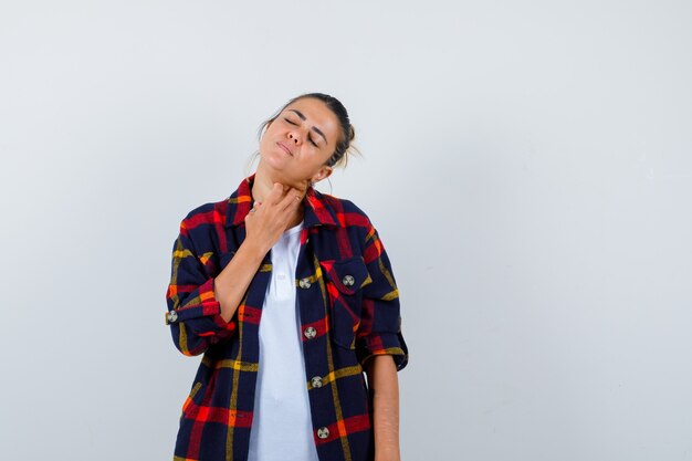 Mujer joven con los dedos en el cuello en camisa a cuadros y luciendo dolorosa, vista frontal.