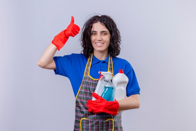 Mujer joven con delantal y guantes de goma con productos de limpieza con una gran sonrisa en la cara mostrando los pulgares hacia arriba sobre la pared blanca