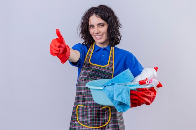 Mujer joven con delantal y guantes de goma que sostienen el lavabo con herramientas de limpieza con una gran sonrisa en la cara mostrando los pulgares hacia arriba sobre la pared blanca