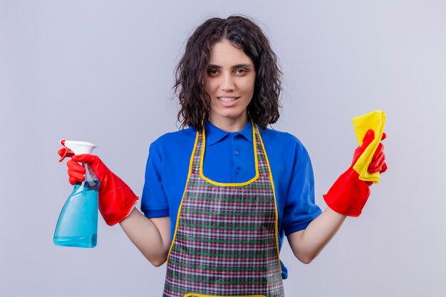 Mujer joven con delantal y guantes de goma sosteniendo una alfombra y spray de limpieza mirando a la cámara con una sonrisa en la cara de pie sobre fondo blanco.