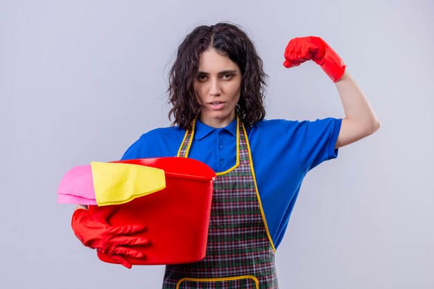 Mujer joven con delantal y guantes de goma sosteniendo el balde con herramientas de limpieza levantando el puño mostrando bíceps mirando confiado, listo para limpiar sobre la pared blanca aislada