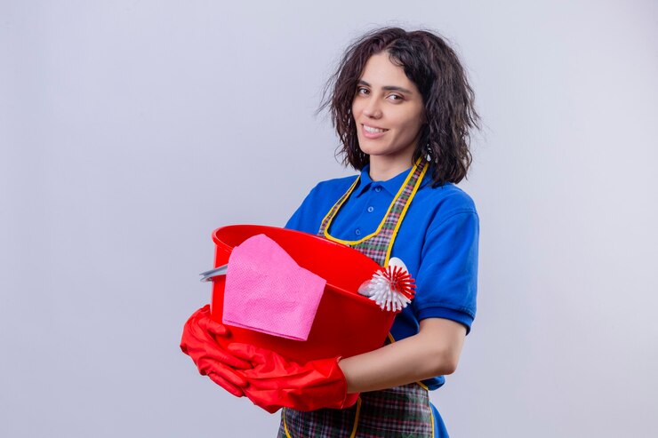 Foto gratuito mujer joven con delantal y guantes de goma sosteniendo un balde con herramientas de limpieza mirando a la cámara con una sonrisa en la cara lista para limpiar de pie sobre fondo blanco.