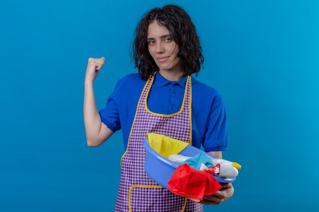 Mujer joven con delantal con lavabo con herramientas de limpieza levantando el puño mostrando bíceps sonriendo confiado, listo para limpiar sobre la pared azul aislada