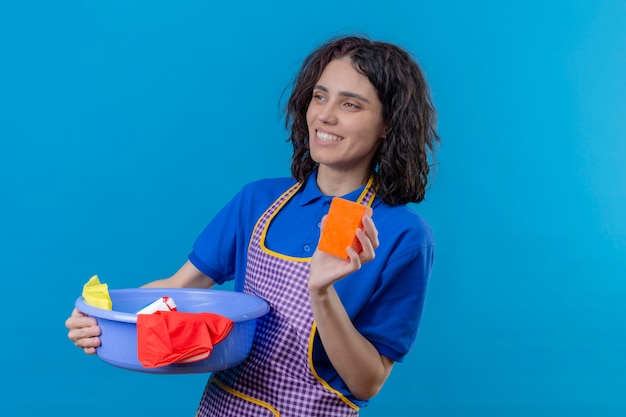 Mujer joven con delantal con lavabo con herramientas de limpieza mostrando esponja en la mano sonriendo alegre mirando a un lado sobre la pared azul