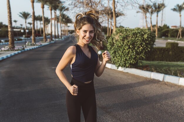 Mujer joven delgada atractiva activa en ropa deportiva corriendo, sonriendo en la calle con palmeras en la ciudad tropical. Modelo de moda, entrenamiento, entrenamiento, estado de ánimo alegre, motivación.