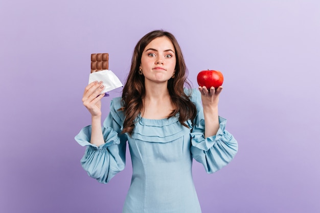 Mujer joven delgada elige entre manzana sana y chocolate dulce. Morena no puede decidir qué comer para el almuerzo.