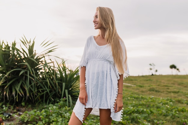 Mujer joven delgada con largo cabello rubio hermoso vestida con vestido blanco posando al atardecer