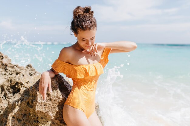 Mujer joven delgada en traje de baño amarillo hermoso mirando hacia abajo mientras posa en la playa. Magnífica chica caucásica tomando el sol en la orilla del mar.