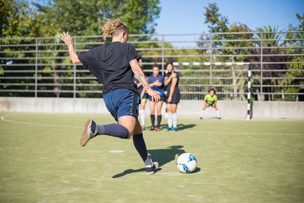 Mujer joven deportiva pateando la pelota en el día de verano. Deportista con uniforme oscuro pateando la pelota en dirección a los goles de fútbol, compañeros de equipo en segundo plano. Deporte, ocio, concepto de estilo de vida activo.